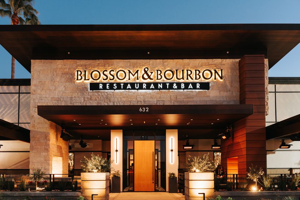 Blossom & Bourbon at dusk, fully finished: halo-lit BLOSSOM & BOURBON letters over the stone entry tower, RESTAURANT & BAR on a slim backer below, warm light spilling from the patio.