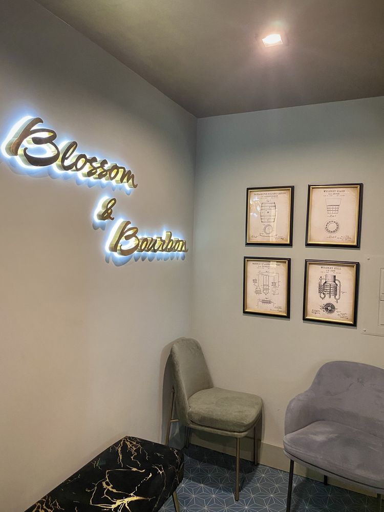 Inside the restaurant: polished script lettering reading Blossom & Bourbon, halo-lit in cool white on the lounge wall beside framed whiskey patent prints.