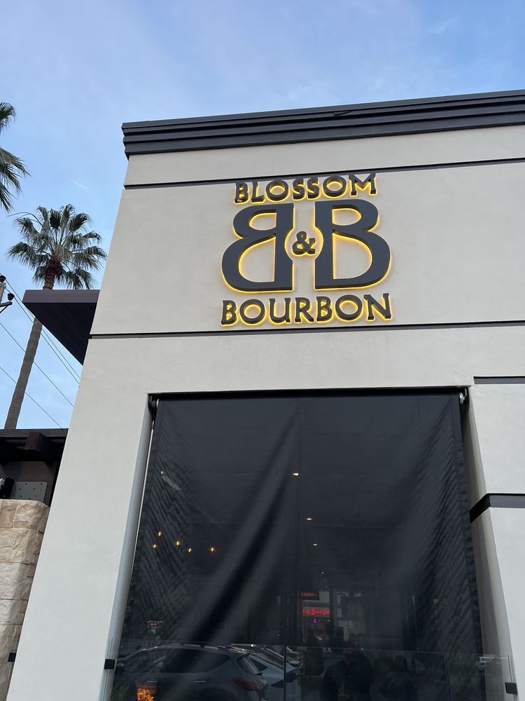 The finished B&B monogram on the side wall at dusk: dark metal letters ringed in warm amber halo light against pale stucco.