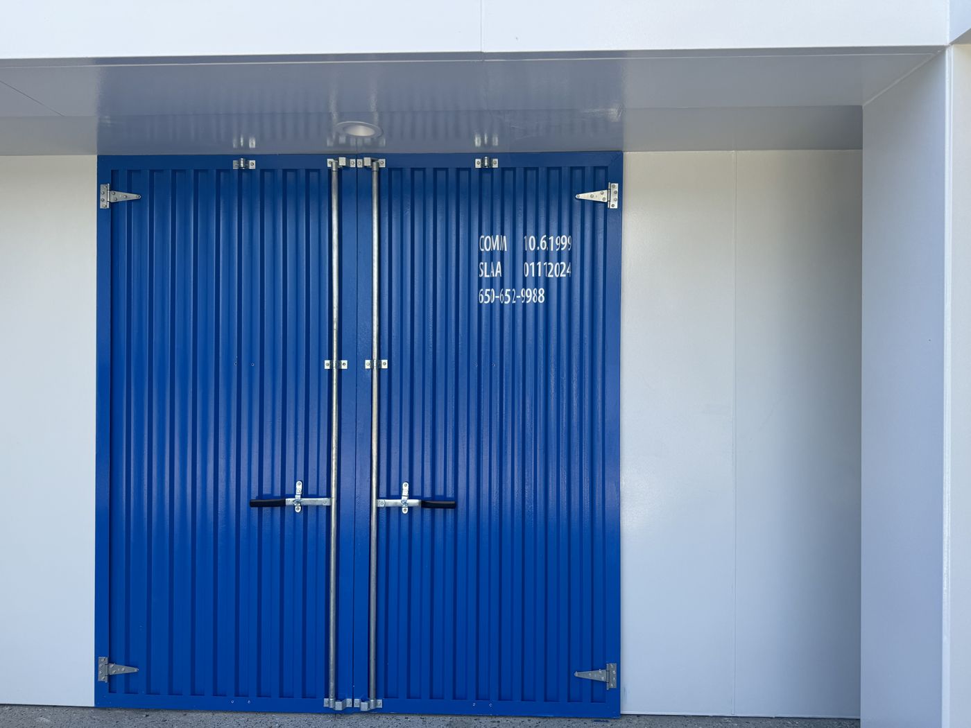 An exterior double door finished as a blue shipping container door with corrugated steel, hinges, lock rods, and stenciled markings.