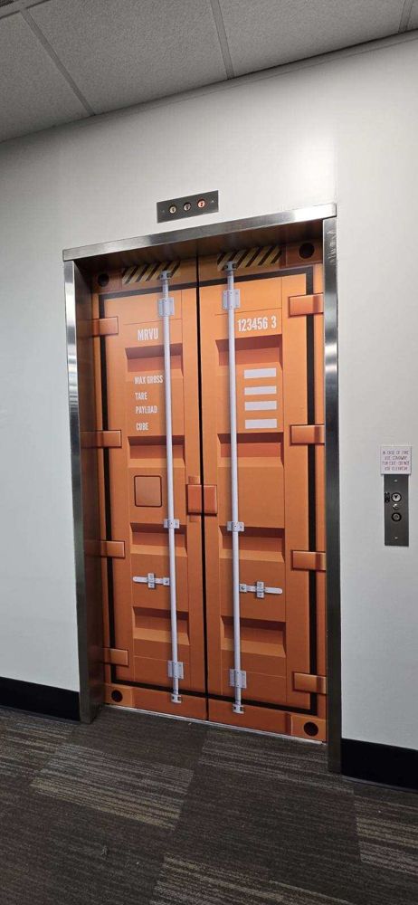 Elevator doors printed as an orange shipping container in a carpeted office corridor.