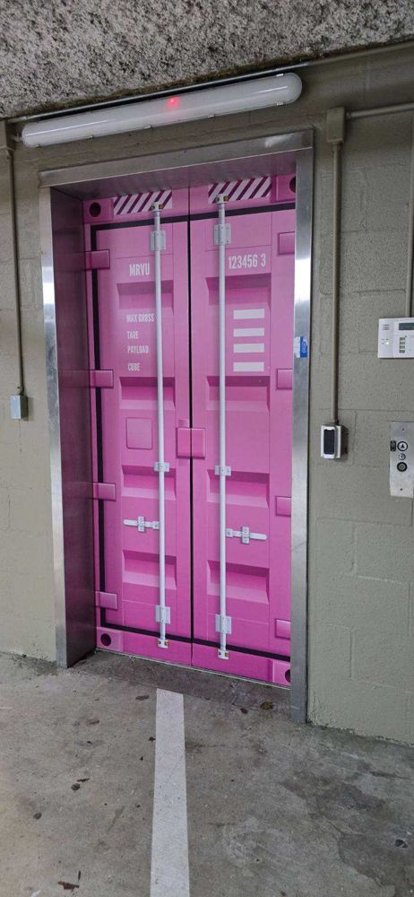 Elevator doors printed as a pink shipping container with white lock rods and stenciled cargo markings, set in a concrete garage wall.
