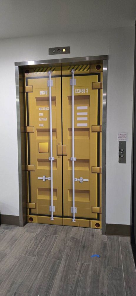 Elevator doors printed as a yellow shipping container, full scale, in an office hallway.