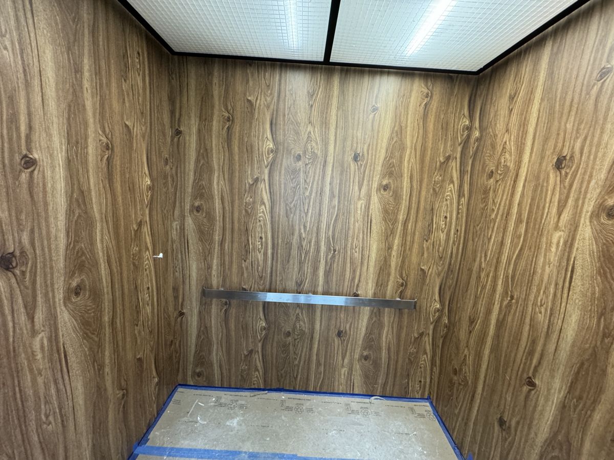 The elevator cab before the wrap: plain wood-grain laminate walls and a stainless handrail.