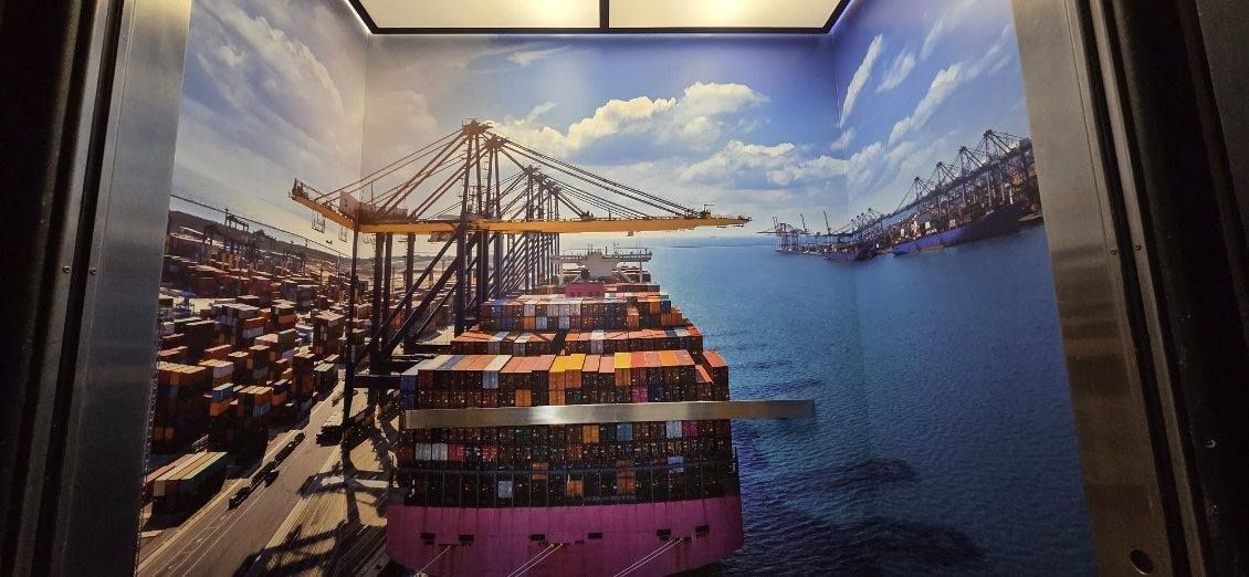 Elevator cab interior wrapped floor to ceiling as a container terminal: gantry cranes over a loaded container ship, stacked containers, open water.