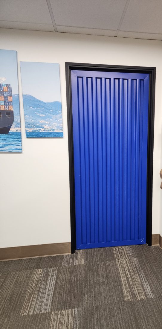 A restroom door fabricated as a blue corrugated container door, flanked by wall murals of a container ship at sea.