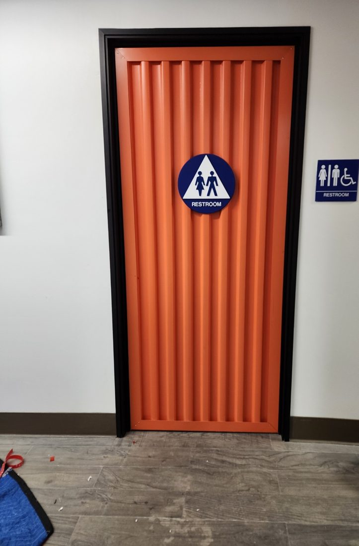 A restroom door fabricated as an orange corrugated container door, with a blue triangle-and-circle restroom sign on the door and an ADA plaque beside it.