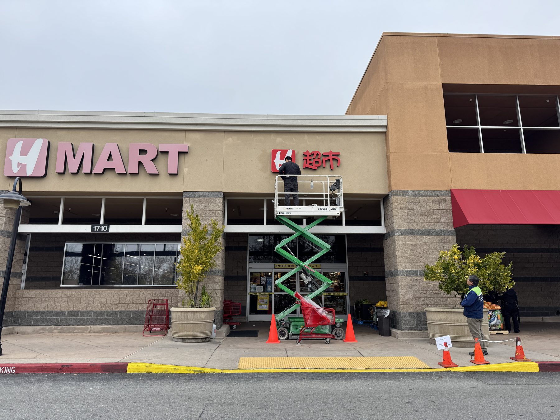 H Mart storefront: on the left, a faded pink H MART sign in aged red acrylic; on the right, a DT Signs installer on a scissor lift mounting a vivid red UV-film sign on the same wall.