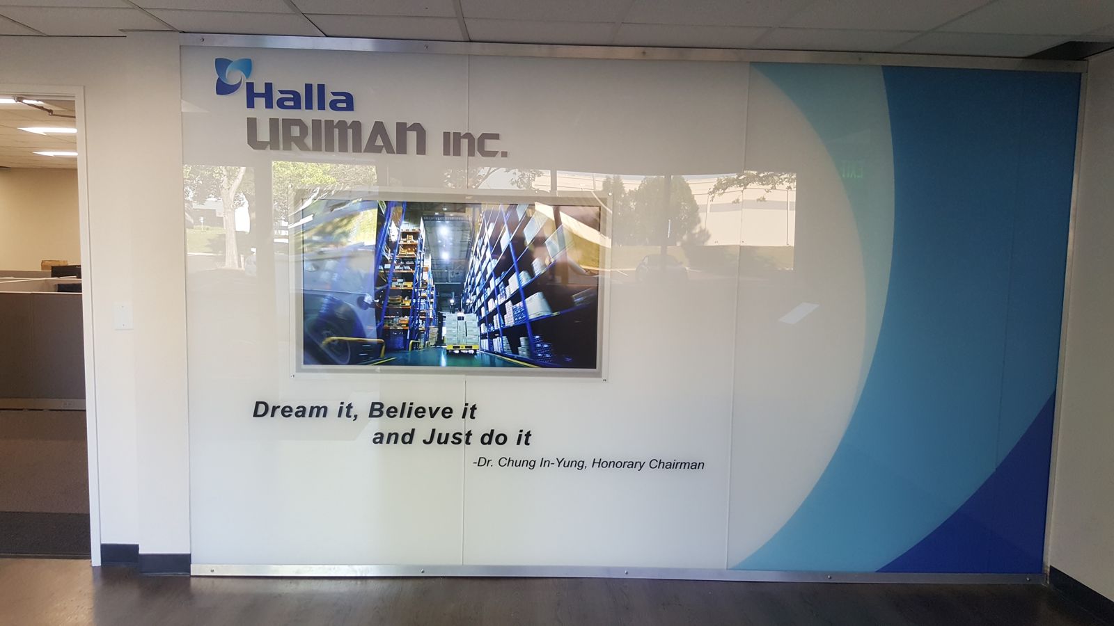 Halla Uriman Inc. lobby feature wall: white brand panel with the Halla logo, a framed display screen, sweeping blue graphic band, and the honorary chairman's quote — Dream it, Believe it and Just do it.