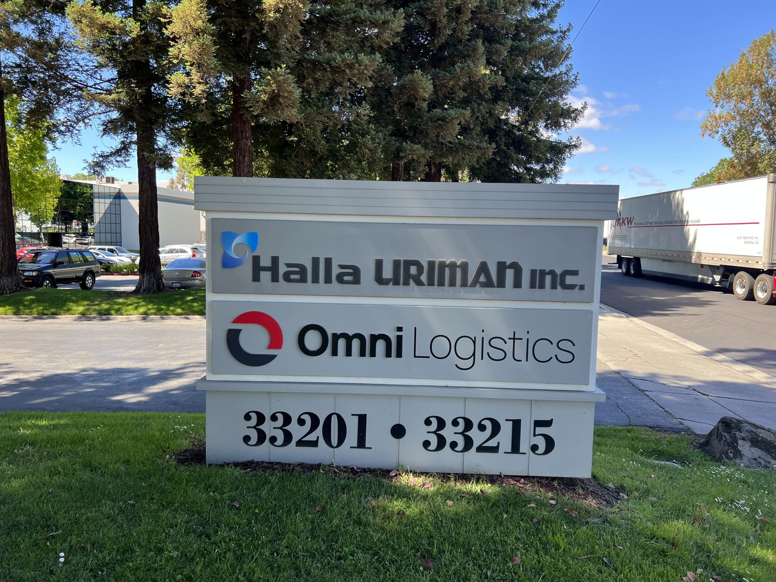 Street monument sign carrying the Halla Uriman Inc. and Omni Logistics logos on stacked panels, set in a lawn with redwoods and a semi truck passing behind.