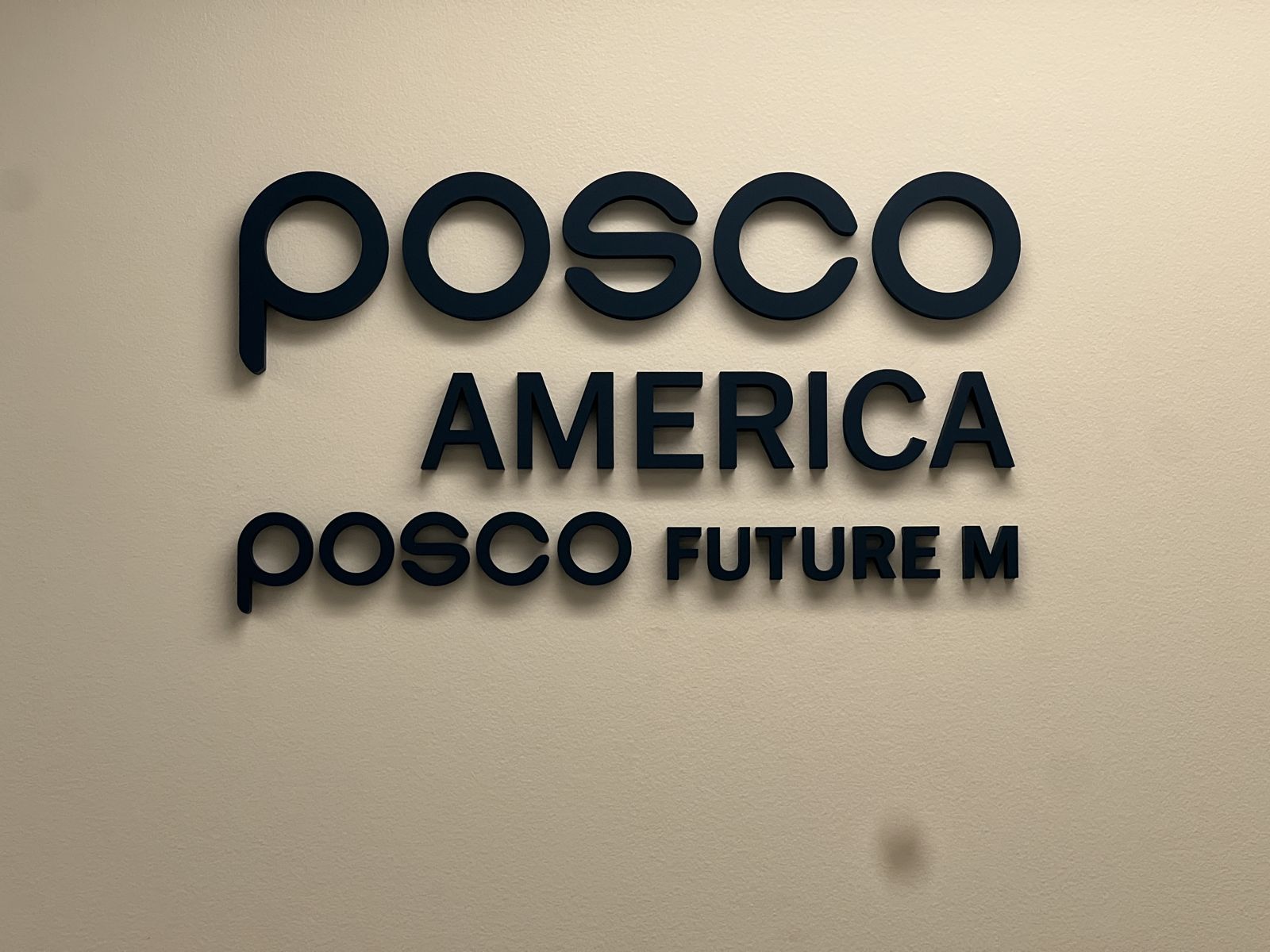 POSCO AMERICA and POSCO FUTURE M in dark dimensional letters, stacked on a warm white interior wall.