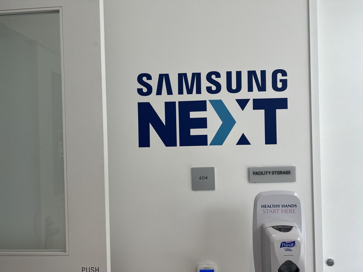 Samsung NEXT logotype in blue dimensional graphics on an interior wall, with an ADA-compliant suite number plaque and facility signage beside it.