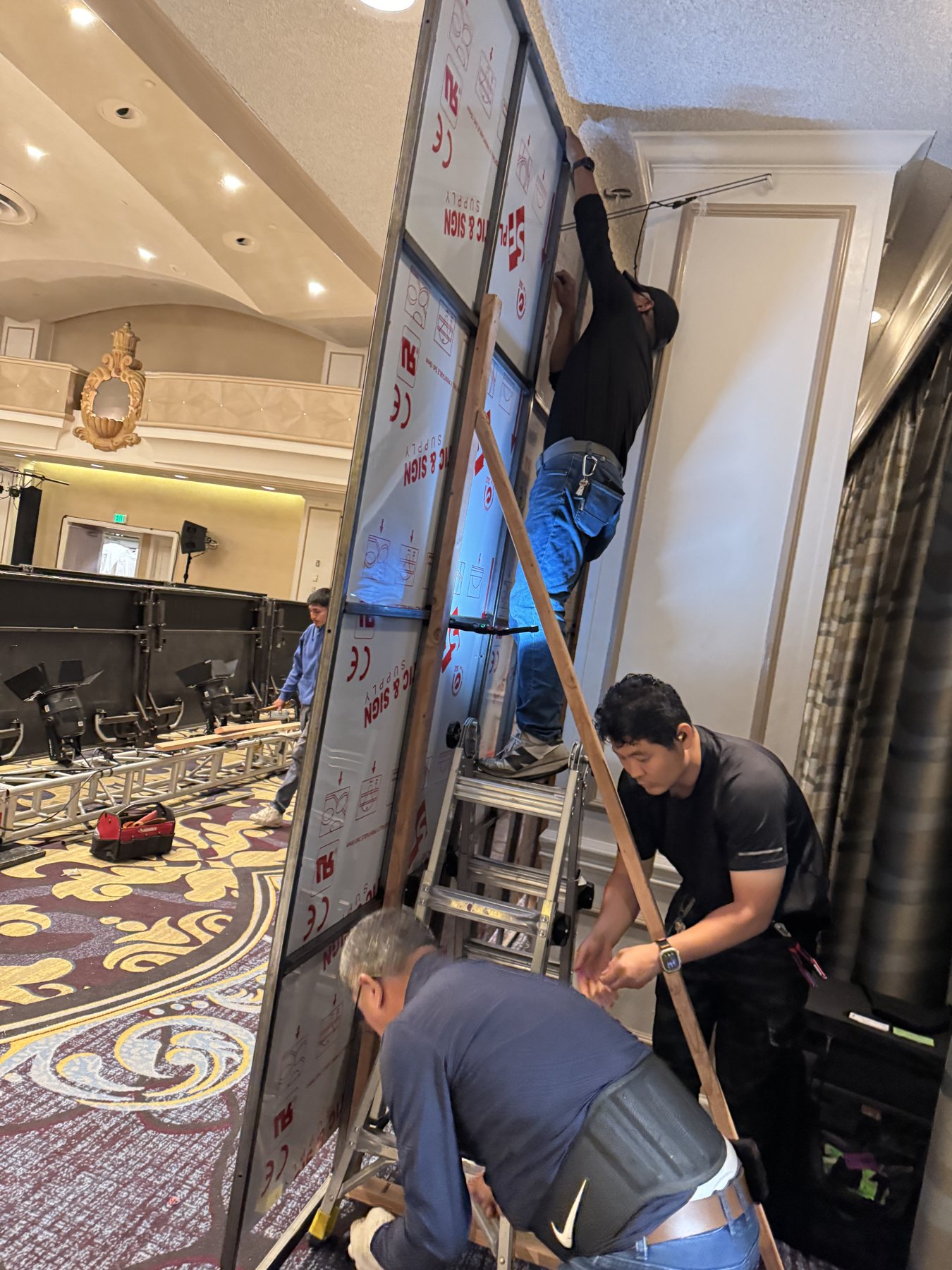 DT Signs crew raising and bolting a steel-framed wall section inside a hotel ballroom.
