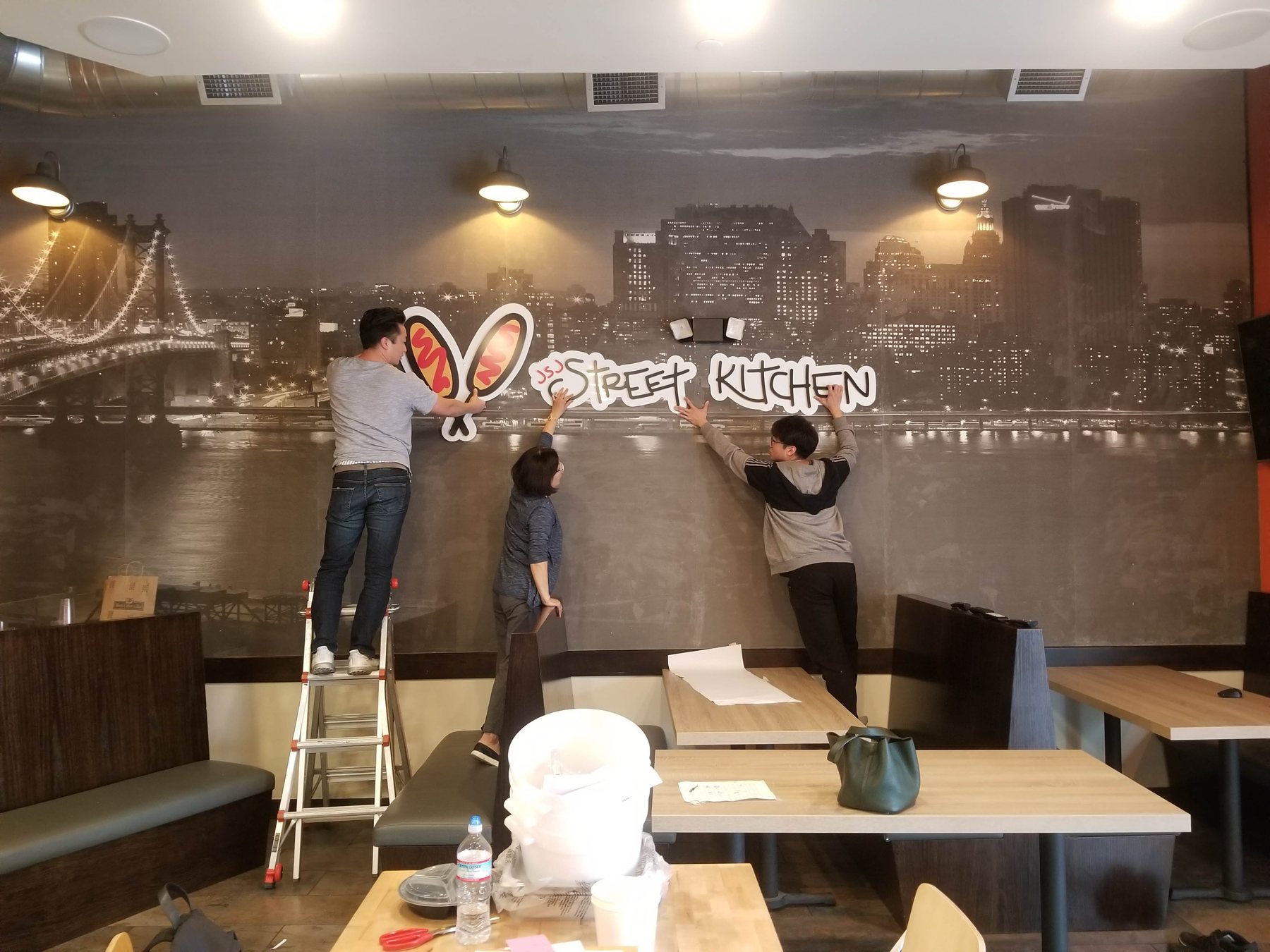 DT Signs crew on ladders finishing a night-city mural wall inside a restaurant dining room.